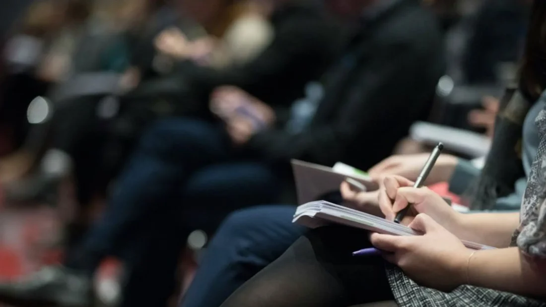 Headerbild für die Termine der GK KFO 2025: Personen in einem Raum hören einen Vortrag an