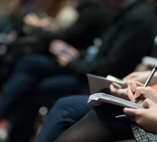 Headerbild für die Termine der GK KFO 2025: Personen in einem Raum hören einen Vortrag an